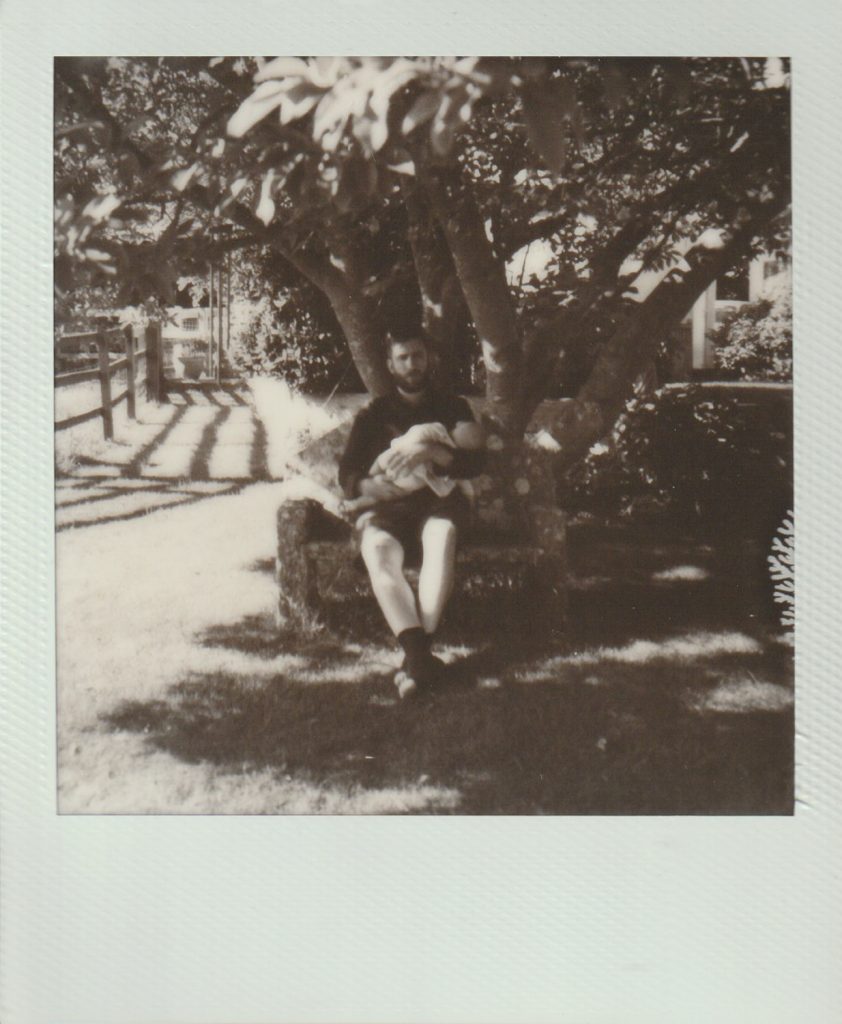 Polaroid film of a father sitting on a bench under a magnolia tree, holding his sleeping daughter.