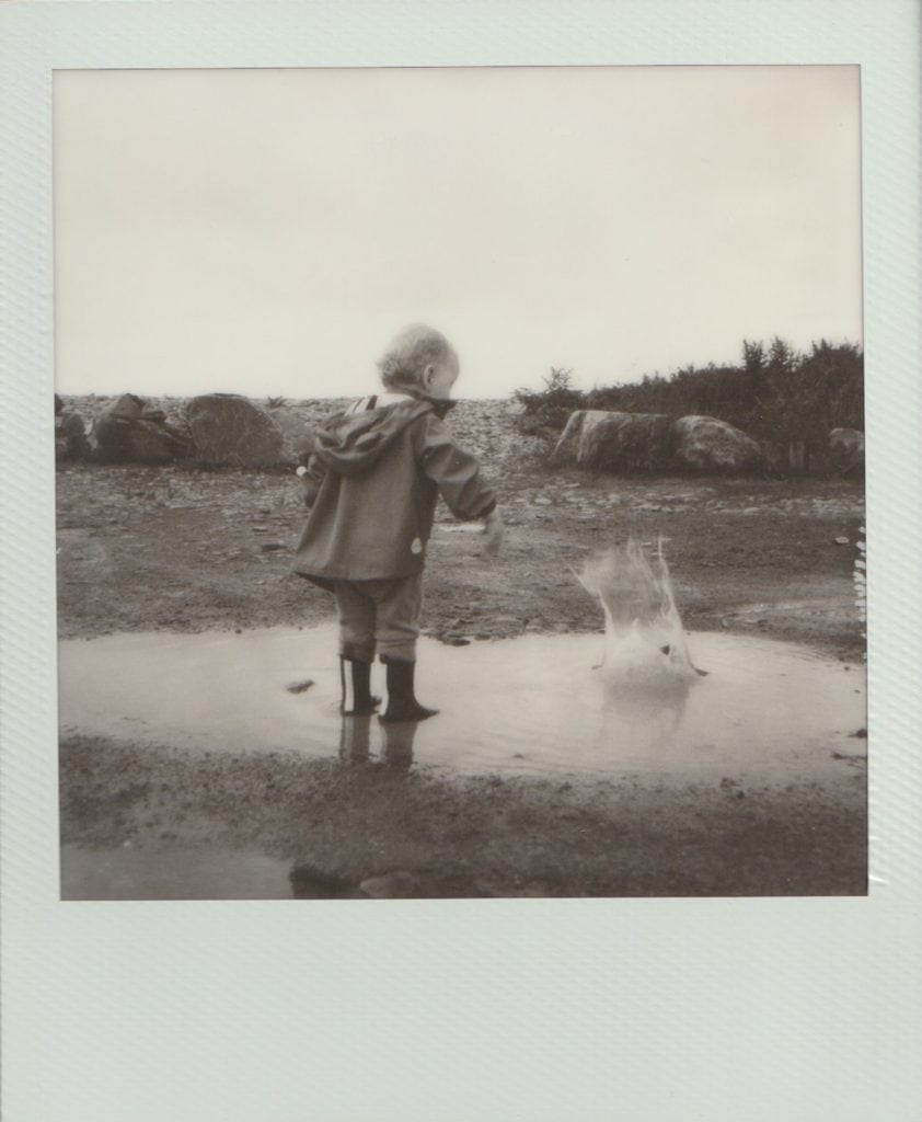 Polaroid film of a small boy throwing a large rock into a puddle, causing a huge splash.