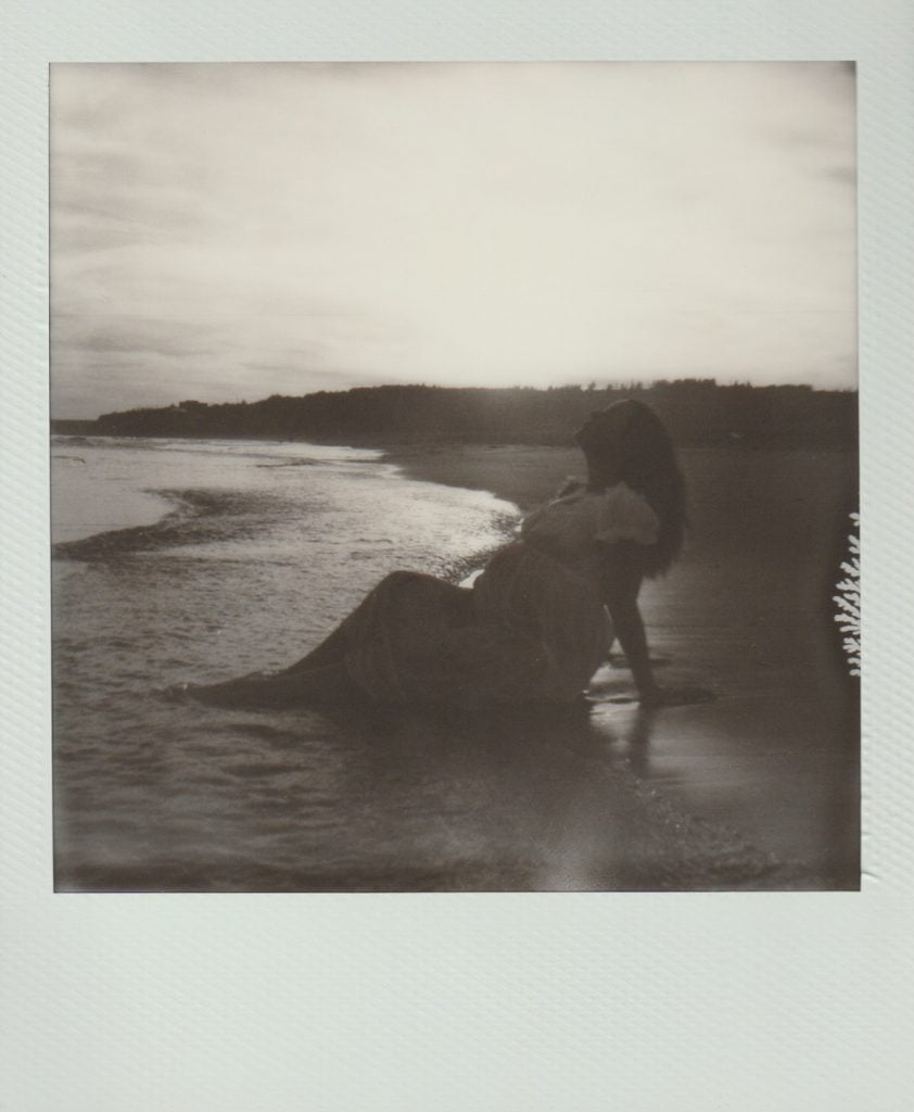 A pregnant woman sits on a beach at sunset and looks to the sky. Polaroid Film.