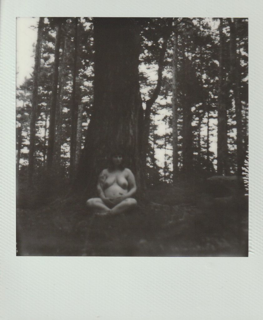 Polaroid film of a pregnant woman sitting naked on the ground, under a tree
