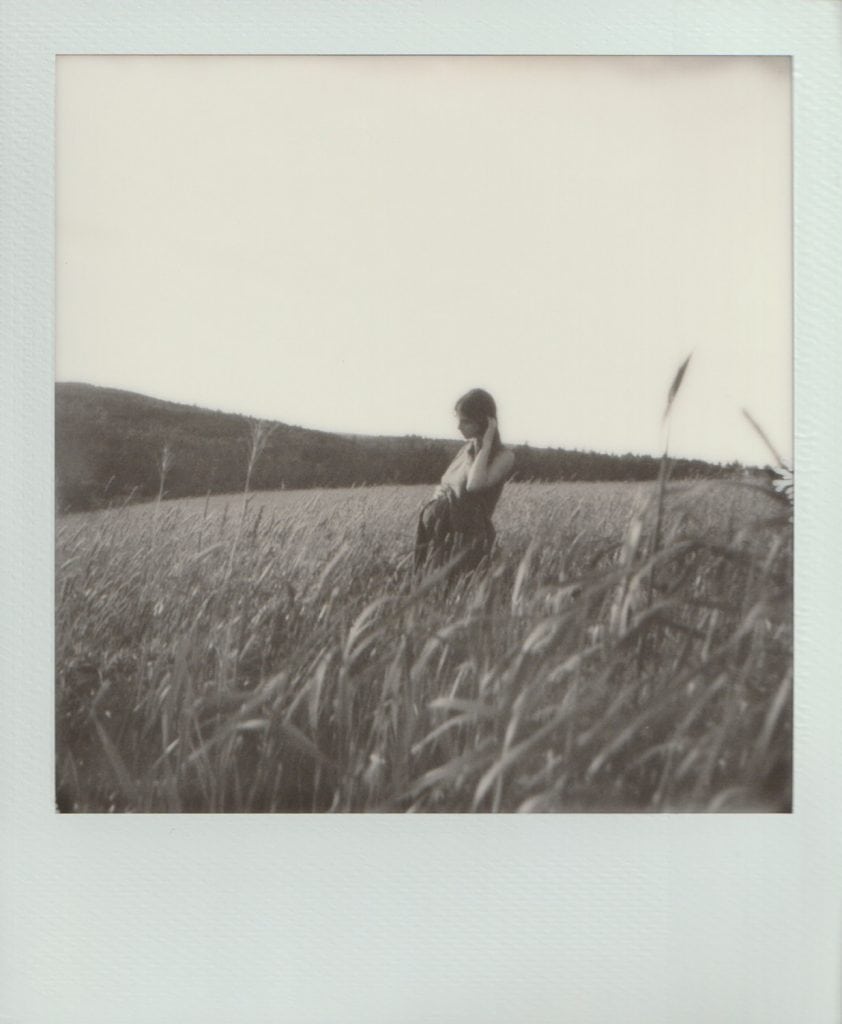 Polaroid film of a pregnant mother tucking her hair behind her ears as she stands in tall grasses.