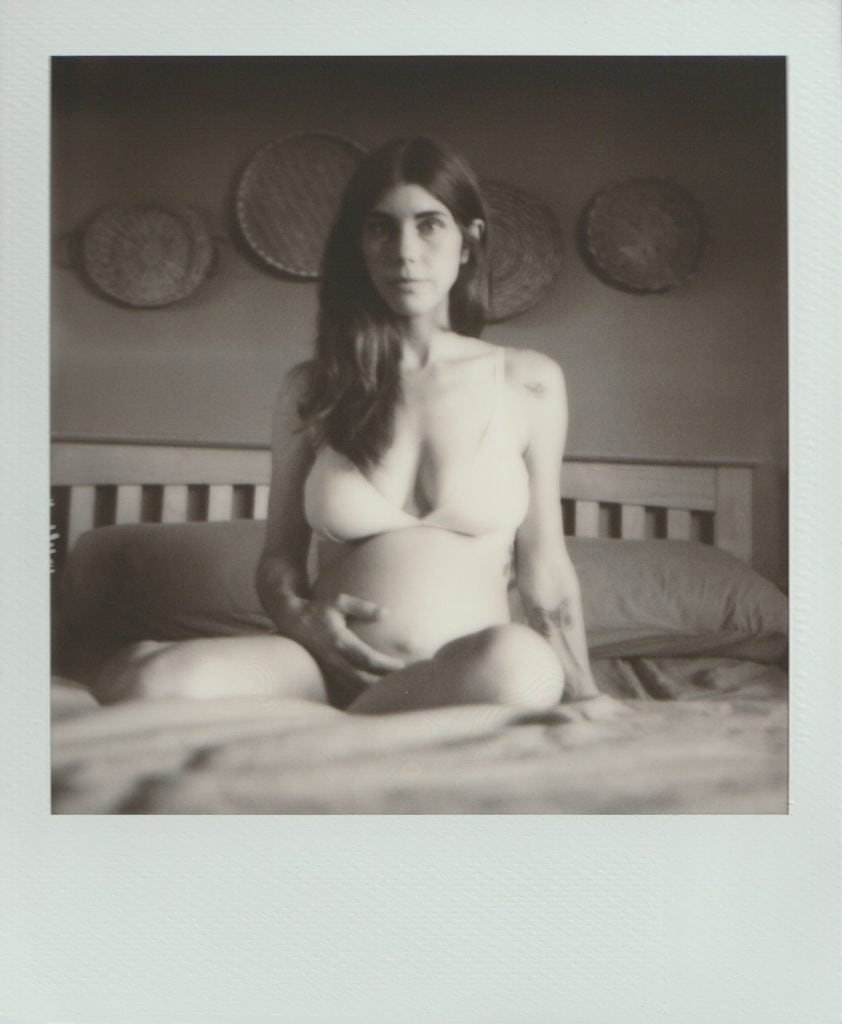 Polaroid film of a pregnant woman sitting on her bed, looking directly at the camera with a hand on her belly.